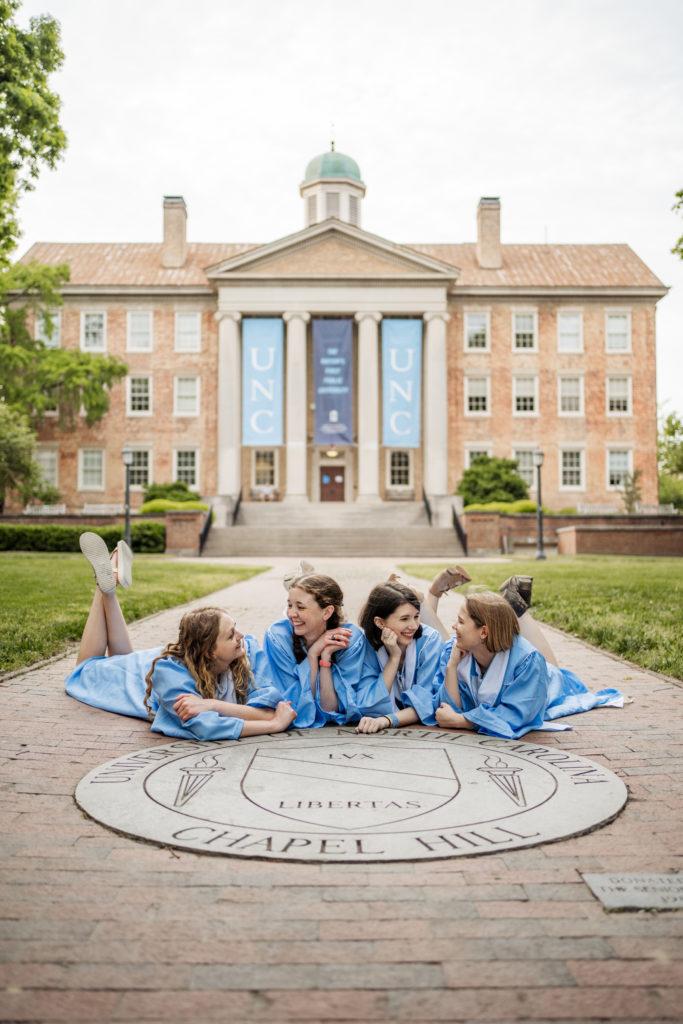 Four UNC students in graduation robes smiling by the seal