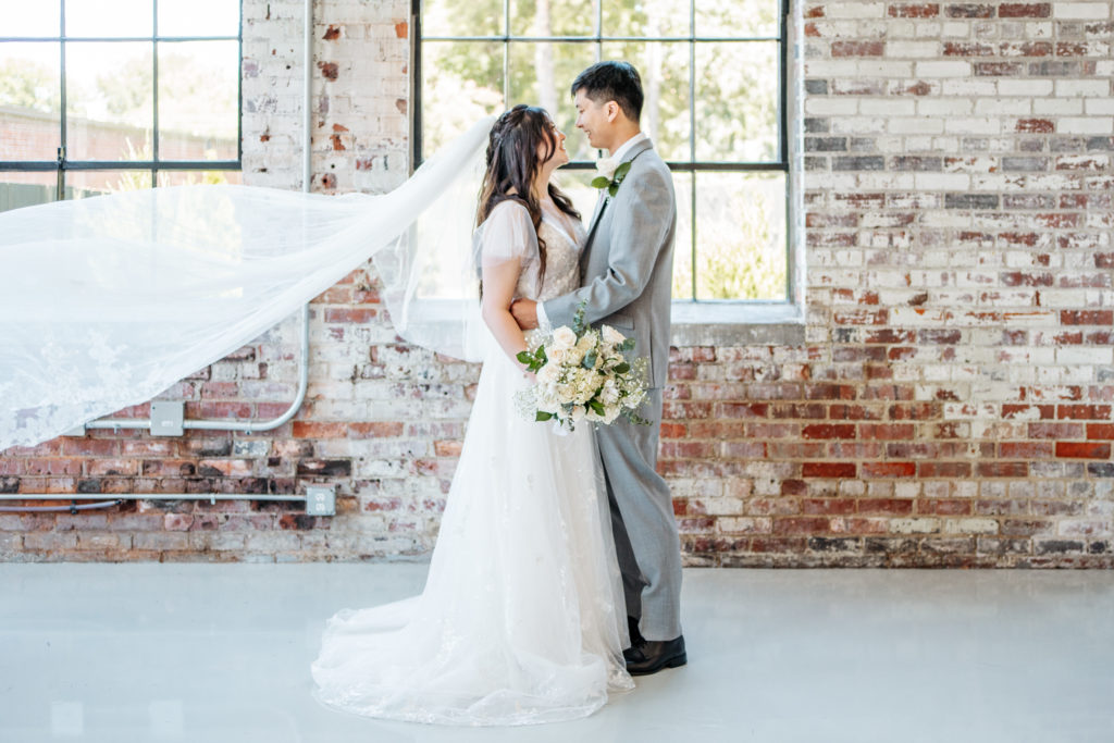veil flutters during bride and groom portrait standing in front of window and brick wall