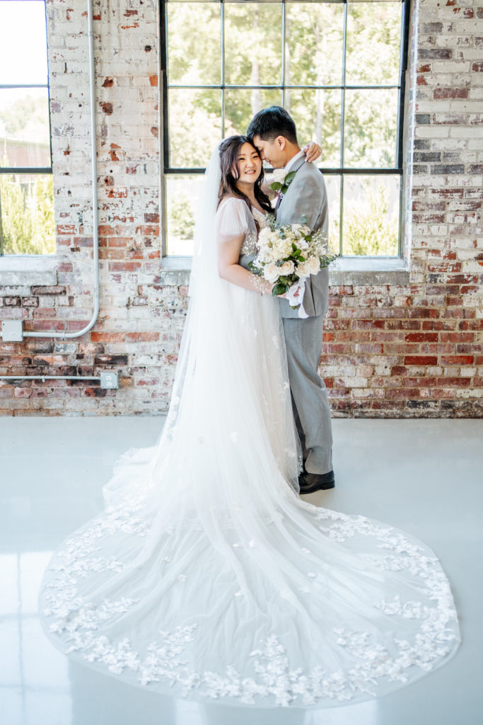 full veil is seen while bride and groom stand together in front of window and brick wall