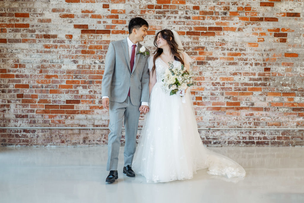 bride and groom holding hands and looking at each other in front of brick wall