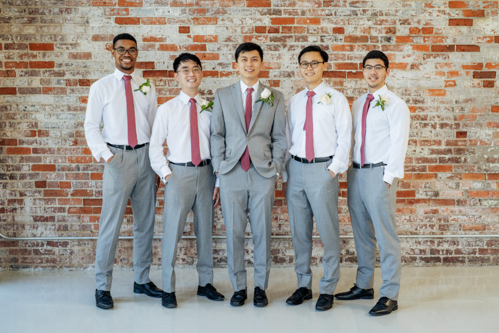 groom stands with groomsmen with their hands in their pockets in front of brick wall