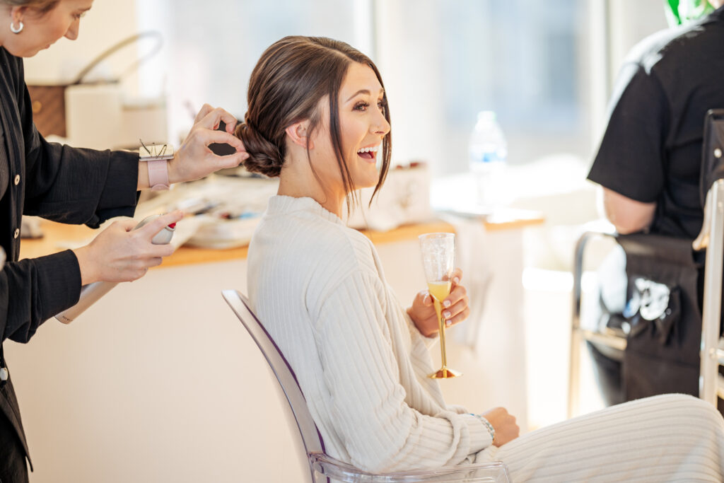 Bride laughing with mimosa while getting her hair done at the Glass Box in Raleigh.
