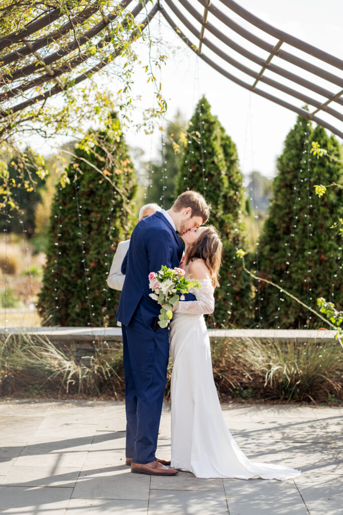 bride and groom first married kiss at wedding ceremony at JC Raulston Arboretum rose garden