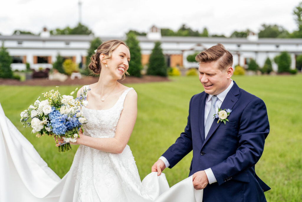bride smiles at the groom as he carries her dress at a wedding at the Evermore near Raleigh